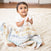 A dark-haired baby in a white onesie sits on a fluffy white rug, holding an aden + anais Disney™ boutique 100% cotton muslin swaddle blanket. The vibrant blanket contrasts with the wooden flooring backdrop as the curious infant gazes upwards.