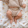 A baby in a beige knitted sweater and shorts lies on a patterned aden + anais boutique 100% cotton muslin swaddle blanket, adorned with colorful trees and rainbows, ensuring comfort while their legs and feet peek out adorably.