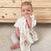 A baby with short hair sits on the carpet, playfully chewing on an aden + anais boutique 100% cotton muslin swaddle. Theyre wearing a white shirt and light-colored pants. In the background, a wooden dresser with round knobs enhances the cozy ambiance.