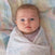 An infant with blue eyes is cozily wrapped in an aden + anais organic cotton muslin swaddle blanket from the 4 pack, showcasing soft, pastel colors and patterns. The GOTS-certified blanket complements the babys calm expression and light brown hair.