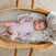 A baby wearing a light purple onesie lies in a wicker basket on an aden + anais organic cotton muslin swaddle blanket from the 2-pack set, atop a soft, patterned surface. The neutral-toned rug complements the scene as the baby gazes wide-eyed with a calm expression.