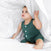 A toddler in a green sleeveless outfit sits under a white patterned aden + anais boutique silky soft swaddle blanket. The setting feels cozy and bright, suggesting a playful bedroom moment with breathable, bamboo-infused viscose for enhanced comfort.