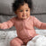 A curly-haired baby sits on a bed, smiling with arms outstretched, in a pink ribbed onesie. Theyre surrounded by aden + anais boutique silky soft swaddle blankets; the bedding features a white floral pattern with a gray cushion in the background.