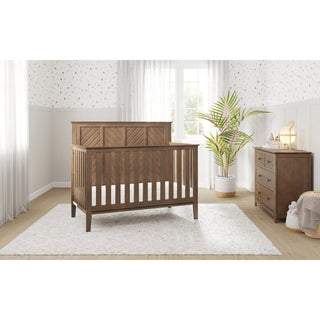 A bright nursery with HALO's Atwood Crib Nursery Bundle, including a convertible crib and matching wooden dresser on a white rug, potted plant, and sunlight through sheer curtains. Light walls feature a subtle dotted pattern.