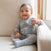 A smiling baby in a gray onesie sits on a light armchair, wrapped in the HALO Fall/Winter Swaddle Bundle, with one arm on the chair and natural light streaming in from a nearby window.