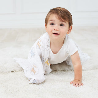 A baby wearing a white onesie crawls joyfully on a soft carpet, clutching a colorful Disney™ essentials cotton muslin swaddle blanket by aden + anais, against a light wall backdrop that enhances this tender moment.