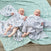 Two smiling babies lie on a light blue patterned blanket outdoors, snug in aden + anais essentials cotton muslin swaddles. One has abstract designs, the other stripes, both made from breathable cotton muslin. Theyre content and comfy under the sky.