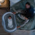 A baby sleeps in the HALO BassiNest® Connected Swivel Sleeper 3.0 smart bassinet with 360° swivel, positioned next to a bed where two adults lie under green blankets. A woman lovingly rests her hand on the bassinet, watching over the baby.