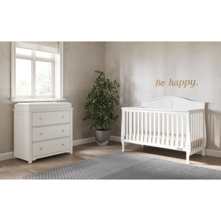 A bright, minimal nursery with a Child Craft Camden 4-in-1 Convertible Crib in Matte White, matching dresser, potted plant, window blinds, gray rug, and “Be happy.” text on a beige wall.