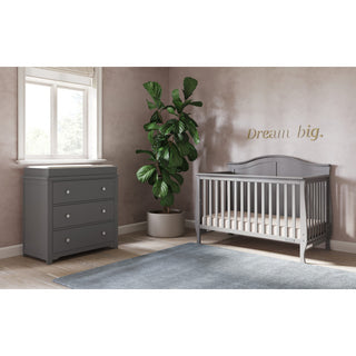 A modern nursery featuring a Child Craft Camden 4-in-1 Convertible Crib in Cool Grey, a matching dresser, potted plant, blue rug, and window. “Dream big.” is written in gold on the wall above the crib.