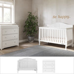 A cozy nursery featuring the Child Craft Camden 2-Piece Nursery Set With Crib and Dresser, plus a potted plant, soft rug on wooden floors, and sunlight from the window. "Be happy." is written above the crib.