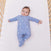 A baby rests in a wooden crib, wearing an aden + anais comfort knit™ footie, cotton-rich and featuring a white moon pattern. With short brown hair and one arm slightly raised, the baby gazes at the camera from a white mattress.