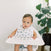 A baby in a high chair wearing an aden + anais boutique 100% cotton muslin snap bib gazes to the side. A large green plant in a pot stands near a white wall behind the baby.