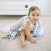 A baby with short brown hair sits on a carpet wrapped in a breathable aden + anais boutique 100% cotton muslin swaddle blanket featuring grey elephants, looking towards the camera with a white cabinet behind.