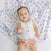 A baby in a white sleeveless top and pink ruffled shorts lies on a breathable, white and blue patterned blanket. Reaching out one hand while looking upward, this little one is as comfy as if wrapped in an aden + anais boutique 100% cotton muslin swaddle blanket.