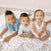 Three young children rest on an aden + anais boutique cotton muslin dream blanket. The older boys have short, styled hair and sport collared shirts in blue and white, while the baby girl wears a white dress and gazes at the camera.