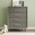 The Child Craft Cranbrook 3 Piece Nursery Set features a modern gray dresser with four drawers and angled legs against a pale green wall. On top are framed photos and books, with a plush teddy bear and wooden blocks nearby.