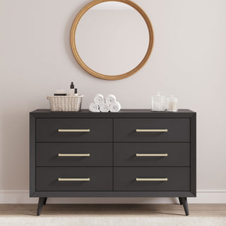 The Child Craft Cranbrook 2 Piece Nursery Set features a sleek black dresser with six drawers against a beige wall, topped with a basket, white towels, and glass containers. A round wood-framed mirror above enhances the mid-century modern appeal.