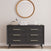 The Child Craft Cranbrook 2 Piece Nursery Set features a sleek black dresser with six drawers against a beige wall, topped with a basket, white towels, and glass containers. A round wood-framed mirror above enhances the mid-century modern appeal.