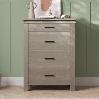 A dresser from the Child Craft Denman 3-Piece Nursery Set with Crib, Dresser and Chest, crafted in light wood finish and featuring four drawers, stands against a green paneled wall as part of a charming nursery furniture collection. Atop the dresser sit a beige pitcher, a white decorative object, stacked books, and an abstract art print while sunlight casts soft shadows across the scene.