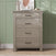 A dresser from the Child Craft Denman 3-Piece Nursery Set with Crib, Dresser and Chest, crafted in light wood finish and featuring four drawers, stands against a green paneled wall as part of a charming nursery furniture collection. Atop the dresser sit a beige pitcher, a white decorative object, stacked books, and an abstract art print while sunlight casts soft shadows across the scene.