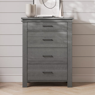The Child Craft Denman 3-Piece Nursery Set with Crib, Dresser, and Chest features a modern gray wooden dresser with four drawers that stands elegantly against a white shiplap wall, blending effortlessly into any stylish nursery. Adorned with decorative items such as a vase and a small sculpture on top, this dresser offers both beauty and functionality for your baby's space.