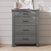 The Child Craft Denman 3-Piece Nursery Set with Crib, Dresser, and Chest features a modern gray wooden dresser with four drawers that stands elegantly against a white shiplap wall, blending effortlessly into any stylish nursery. Adorned with decorative items such as a vase and a small sculpture on top, this dresser offers both beauty and functionality for your baby's space.