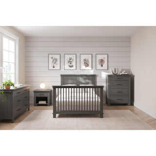 A modern nursery featuring the Child Craft Denman 4-in-1 Convertible Crib in Midnight Gray, with matching gray furniture. Neutral decor, botanical art, and large windows create a calm, cozy craftsman vibe.