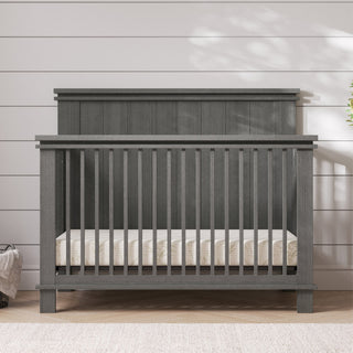 The Child Craft Denman 4-in-1 Convertible Crib in Midnight Gray, featuring vertical slats and a solid headboard, stands on a light rug against a white shiplap wall with a plant partly shown on the right, blending craftsman style and versatility.