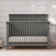 The Child Craft Denman 4-in-1 Convertible Crib in Midnight Gray, featuring vertical slats and a solid headboard, stands on a light rug against a white shiplap wall with a plant partly shown on the right, blending craftsman style and versatility.