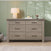 The Child Craft Denman 3-Piece Nursery Set, featuring a spacious dresser with six drawers, is styled against a green paneled wall and topped with a lamp, books, bird-shaped decor, and a small figurine. With a cozy rug and pillow peeking into the scene, this set makes an inviting addition to your baby's nursery.