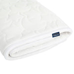 A rolled-up, white quilted DreamWeave Crib Cover by HALO features a textured, hypoallergenic design with a small HALO tag. This breathable mattress pad looks soft and cushioned, ensuring comfortable sleep.