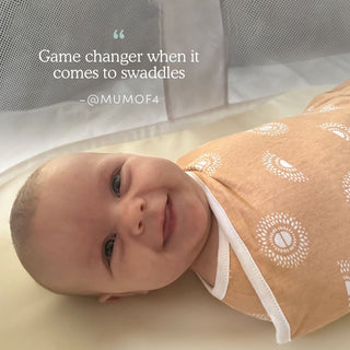 A smiling baby lies in a light brown swaddle from the aden + anais essentials easy swaddle wrap 3 pack, featuring a white pattern. The text above says, Game changer when it comes to swaddle wraps – @MUMOF4. A mesh crib is in the background, ensuring comfort and preventing startle reflex.