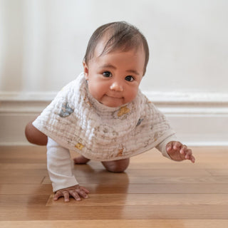 A baby in a white outfit crawls on a wooden floor, smiling and reaching forward while wearing the aden + anais Disney™ Winnie the Pooh essentials cotton muslin burpy bib.