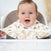A smiling baby with blue eyes and light brown hair sits in a high chair, wearing an aden + anais essentials cotton muslin burpy bib adorned with animal prints, including giraffes and elephants. The softly blurred background highlights the babys joyful expression.