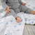 A baby in a gray outfit sits on a white blanket with blue animal prints. The babys legs rest on aden + anais essentials cotton muslin swaddle blanket, while the background shows a textured beige surface.