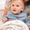 A blue-eyed, light-haired baby, wearing a light blue ribbed shirt with buttons, snuggles on a lap in an aden + anais Disney™ essentials cotton muslin dream blanket featuring animal and tree patterns. The background is softly blurred.