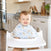 A smiling toddler sits in a high chair in a modern kitchen, wearing an aden + anais essentials cotton muslin burpy bib with animal prints. The countertop and cabinets are visible in the background.