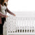 A pregnant woman stands by a white crib, adjusting a patterned baby blanket. Wearing a gray top and mauve cardigan, she delicately arranges the aden + anais essentials cotton muslin crib sheet. The bright room suggests a nursery setting.