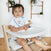 A dark-haired toddler sits on a wooden high chair with a white tray, wearing an aden + anais essentials cotton muslin bandana bib from the 2-pack, featuring colorful patterns. The white wall and hanging plants create a charming background.