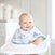 A smiling baby in a white high chair wears an aden + anais essentials cotton muslin burpy bib. The background shows a bright, modern kitchen with a large window and white jars on the countertop.