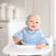 A smiling baby in a high chair wears an aden + anais essentials cotton muslin burpy bib, featuring a cloud pattern. They hold a spoon with white kitchen cabinets and some jars in the background.