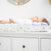 A baby in white lies on an aden + anais essentials cotton muslin changing pad cover, adorned with a colorful dinosaur print, atop a white dresser. The baby looks up while a round mirror is partially visible on the wall above.