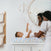 A woman gently changes a babys diaper on an aden + anais essentials cotton muslin changing pad cover with a sea theme. The baby reaches up, and the scene is reflected in a wall-mounted mirror with a mobile hanging above in the cozy, well-lit room.