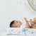 A baby lies on a soft aden + anais essentials cotton muslin changing pad cover, gazing at a mobile with beads, while an adults hands gently support them. The cover features playful bears, suns, and crescent moons. A round mirror is partially visible on the wall.