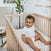 A smiling baby in a white onesie sits in a wooden crib on an aden + anais essentials cotton muslin crib sheet with playful nursery prints. In the background, there’s a potted plant, an armchair, and part of a wall adorned with framed pictures.