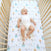 A baby in a white onesie lies on an aden + anais essentials cotton muslin crib sheet adorned with whimsical nursery prints of animals and moons. The wooden-slat crib cradles the smiling baby looking upward.