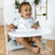 A smiling toddler with curly hair wears an aden + anais essentials cotton muslin snap bib featuring whale designs while sitting in a high chair, holding a white tray. The wooden-floored room and potted plants perfectly capture moments that grow with your babe.