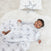 The baby lies on a soft, white rug, wrapped in a Disney™ essentials cotton muslin swaddle blanket by aden + anais with playful patterns. Dressed in gray and smiling, a decorative pillow with a star design is visible in the background.