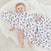 A baby lies on a fluffy white rug, partially covered with an aden + anais Disney™ essentials muslin swaddle blanket featuring colorful cartoon mice. The babys eyes are open, with one arm resting on the breathable cotton blanket.